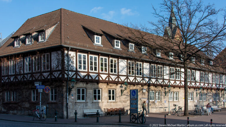 Long half-timbered building with white infill, stone ground floor, red-tiled roof, dormers, and bicycles outside.