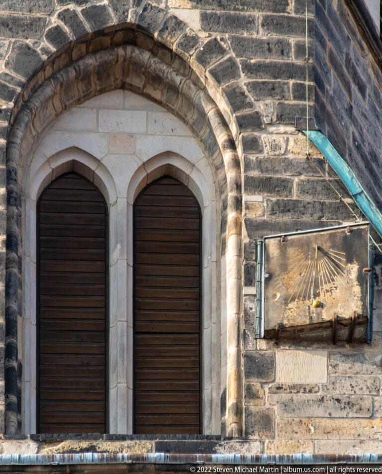 Two shuttered arched openings in stone masonry beside a small wall-mounted sundial.