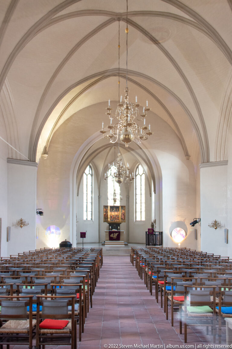 Long church nave with ribbed vault, rows of wooden seating, hanging chandeliers, and an altar at the far end.