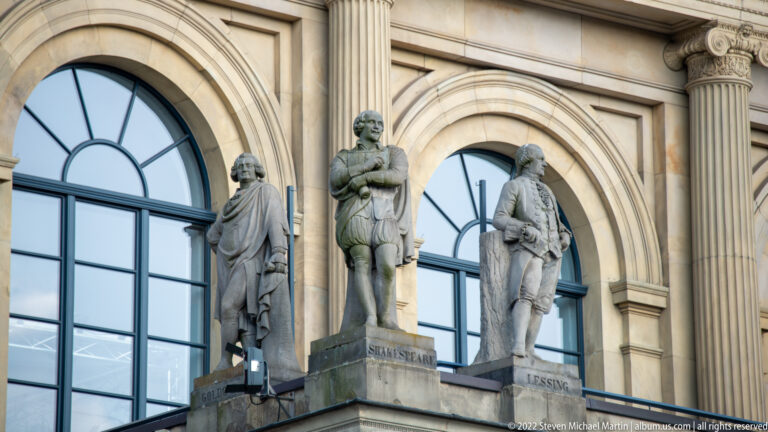 Stone statues between arched opera-house windows, with bases labeled Schiller and Lessing.