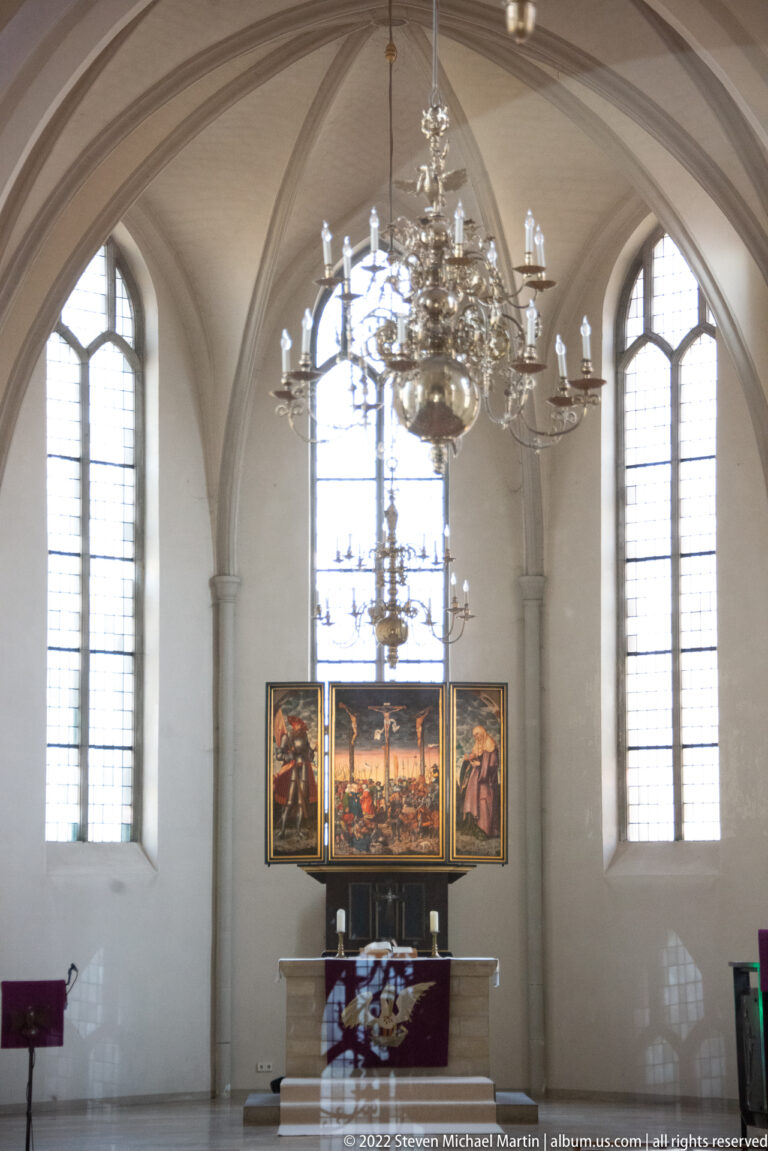 Church altar beneath a painted triptych, flanked by tall arched windows and overlapped by a large chandelier.