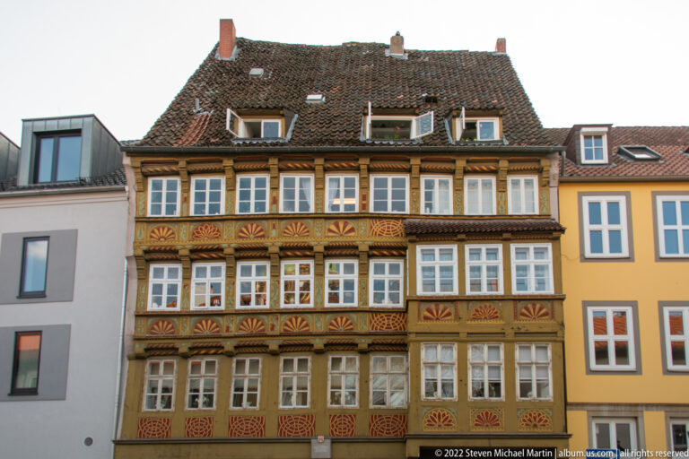 Multi-story timber-framed house with many white windows and richly painted carved panels between floors.