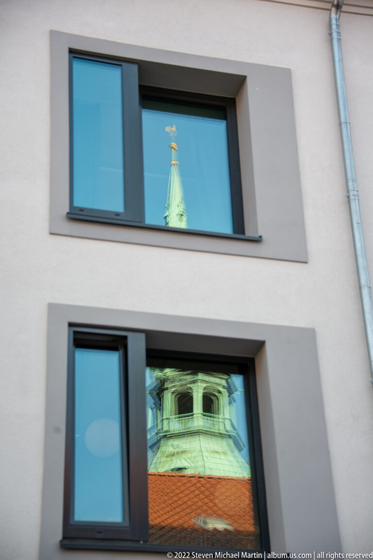 Historic church spire and tower reflected in two rectangular windows of a modern light-colored building.