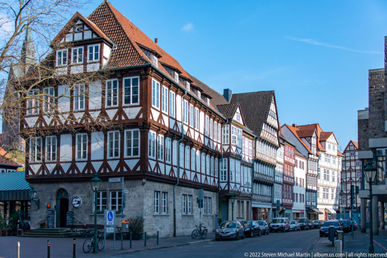 Row of half-timbered old-town buildings lining a narrow street with parked cars, bicycles, and shops.