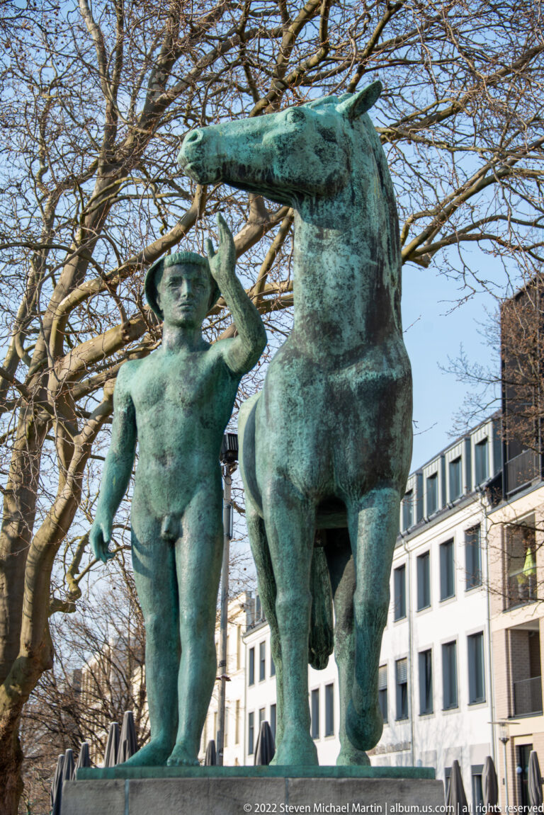 Green-patinated bronze sculpture of a nude child-like figure standing beside a horse.