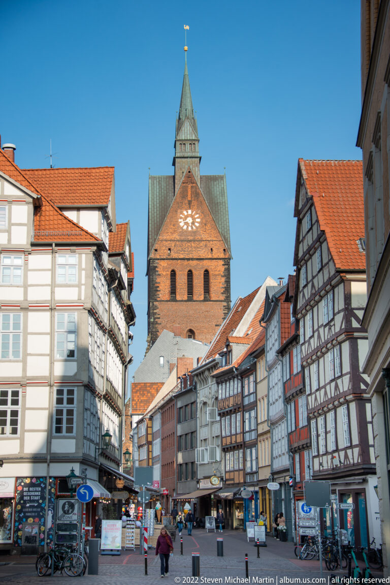 Old-town pedestrian street lined with half-timbered buildings and centered on a brick church tower in the distance.