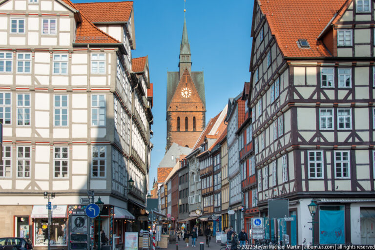 Half-timbered buildings line a narrow street leading toward a brick church tower under a clear blue sky.
