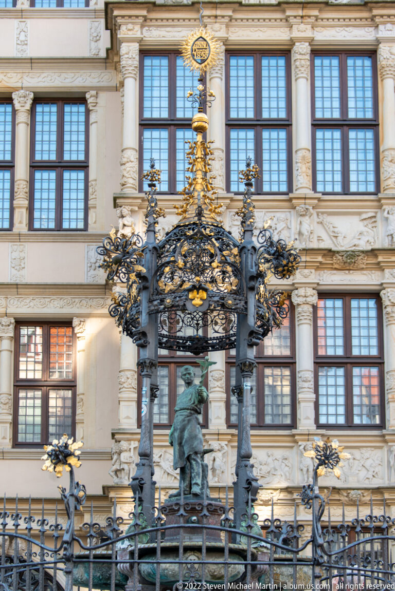 Ornate dark-metal Oskar-Winter-Brunnen with gilded details standing before the decorated façade of the Leibnizhaus.