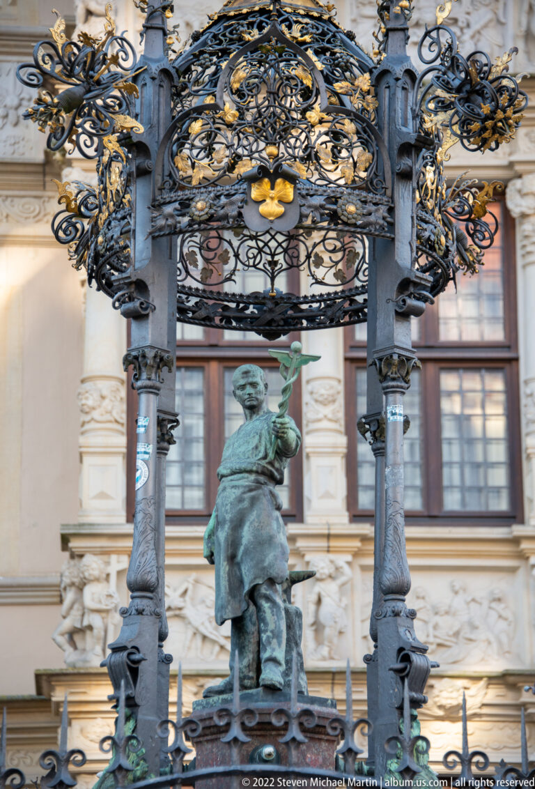Green-patinated standing figure beneath an ornate black metal canopy with gilded details in front of a carved stone façade.