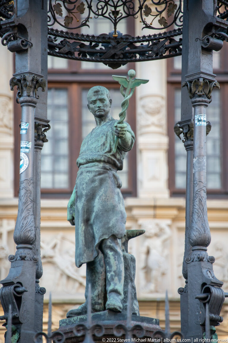 Green-patinated male figure in a long coat holding a leafy branch beneath carved metal supports.