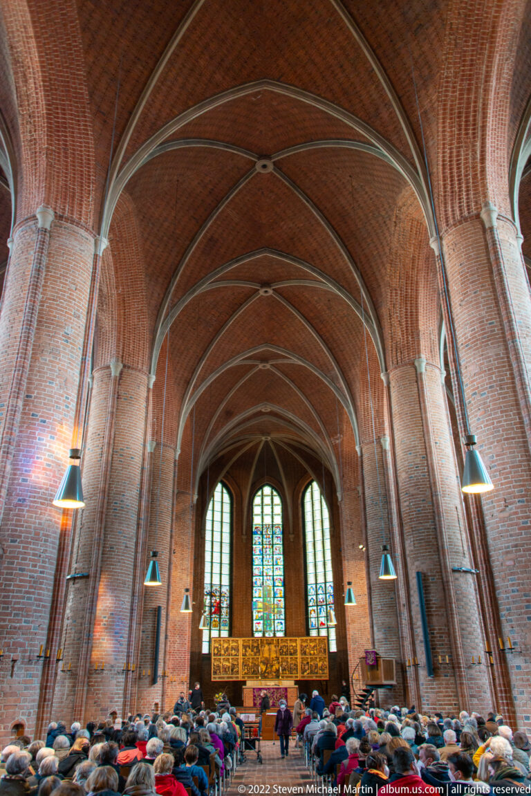 Brick church nave filled with seated visitors beneath high rib vaults, with stained glass and an altar at the far end.