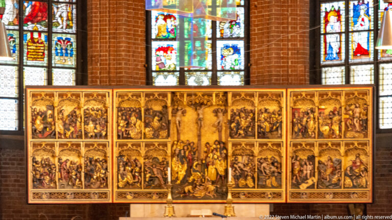 Large gilded multi-panel altarpiece filled with religious scenes, set before stained-glass windows in a brick church.