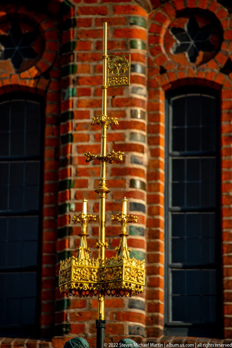 Gilded cross-shaped metal ornament with elaborate openwork standing before dark arched windows in a brick façade.