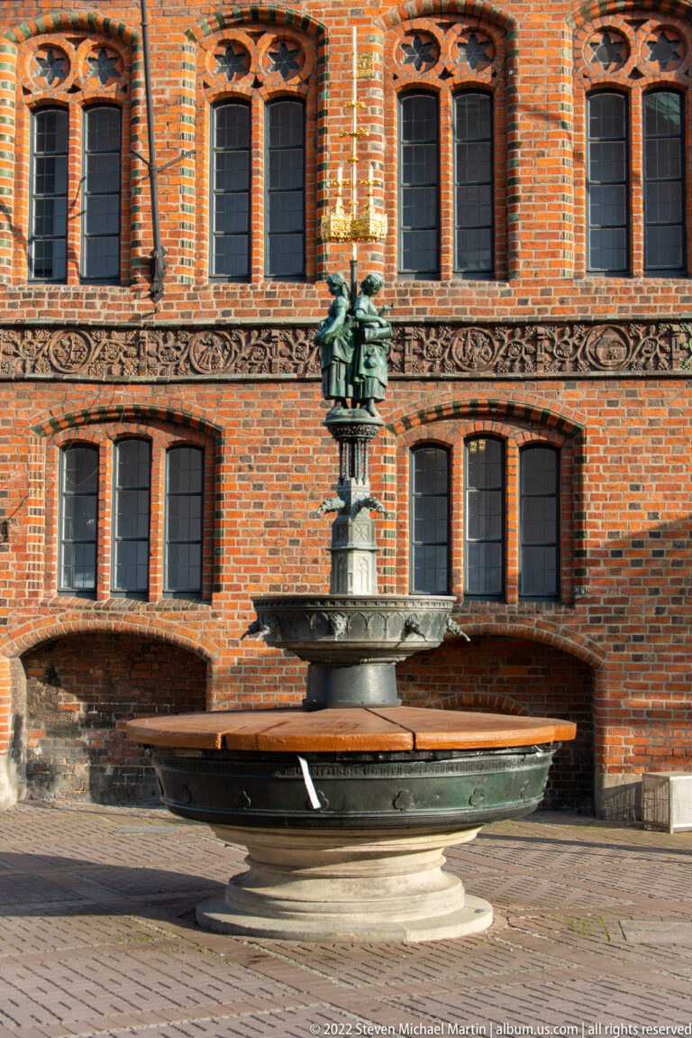 Circular stone fountain basin with a small bronze central figure in front of an ornate red-brick façade.