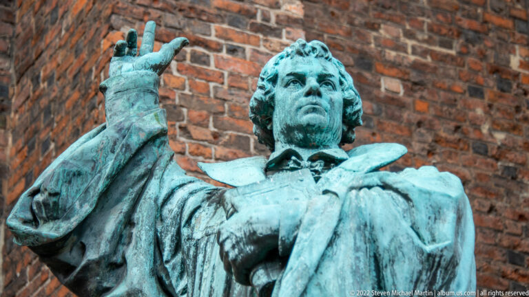 Green-patinated bronze statue of a robed man raising one hand, seen close against a brick wall.