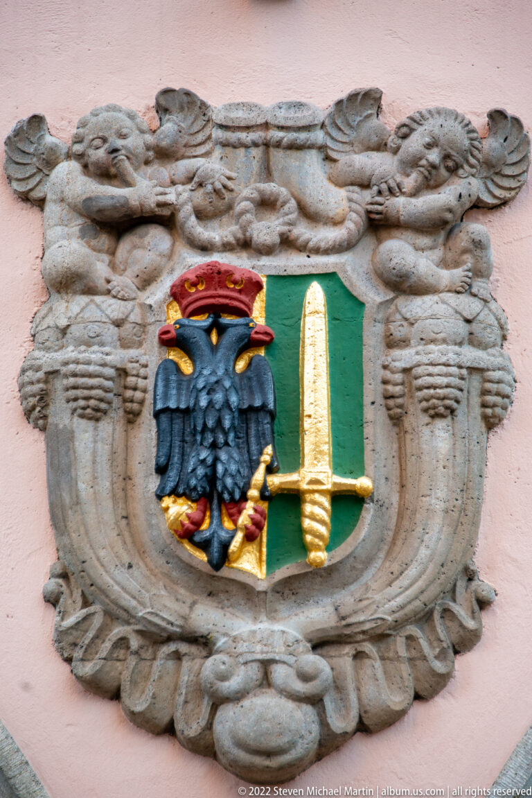 Stone heraldic cartouche with a crowned black eagle and a gold upright sword on a green field.
