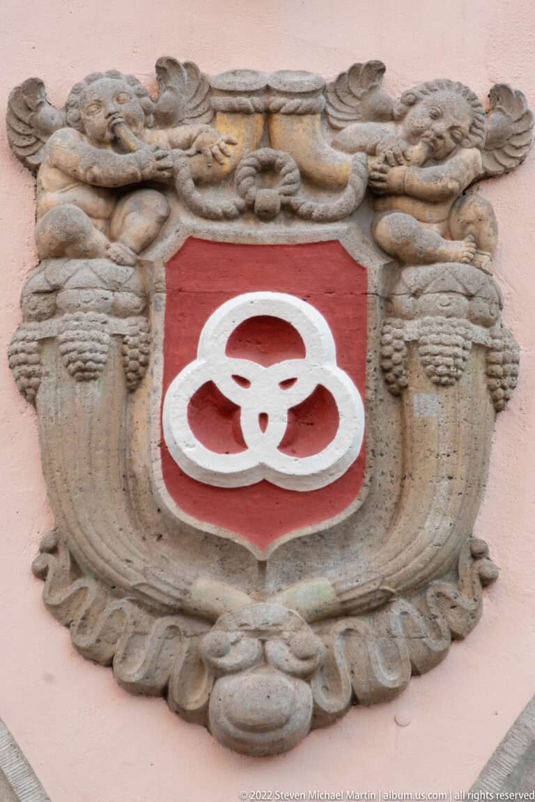 Red heraldic shield with a white three-lobed emblem set within an elaborate carved stone cartouche.