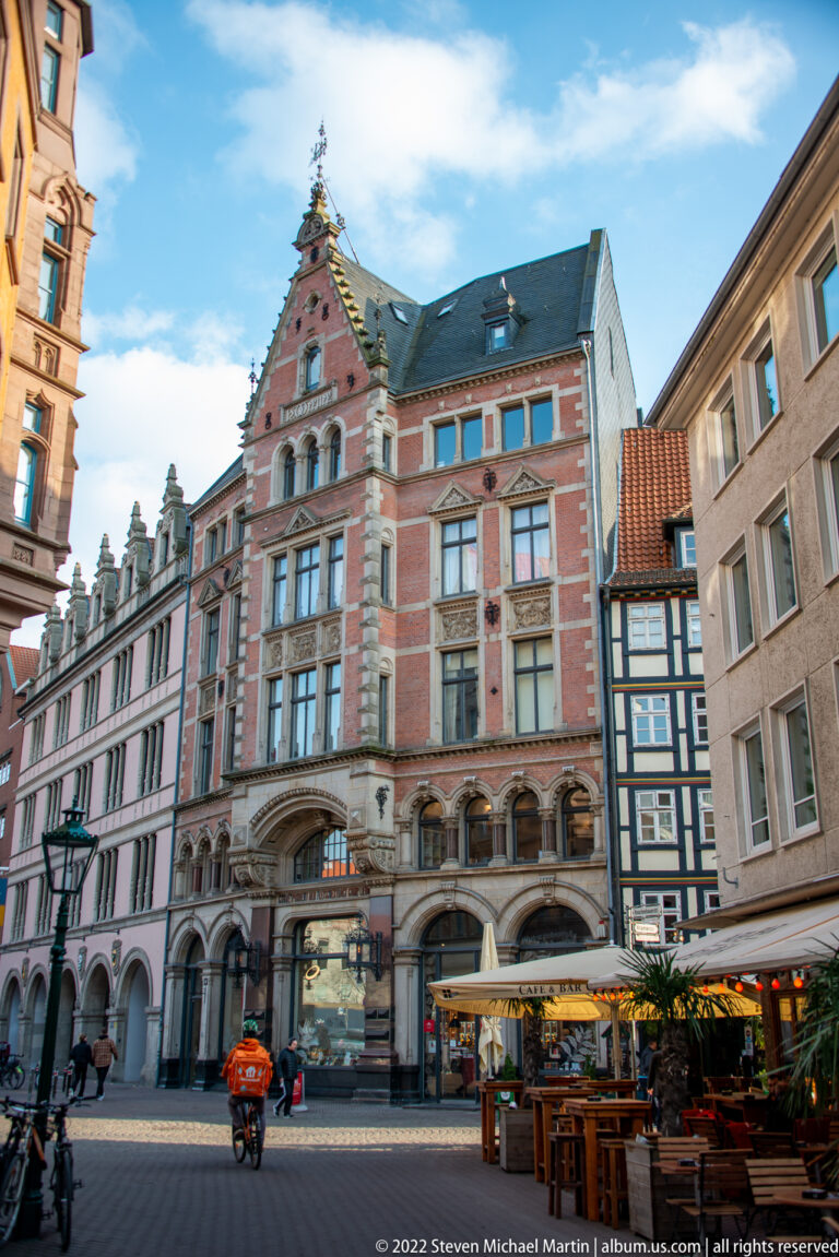 Ornate historic corner building with arched ground-floor openings, tall windows, gabled roof, café seating, and cyclists.