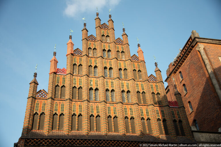 Tall ornate Brick Gothic stepped gable with patterned masonry, narrow arches, and pinnacles against blue sky.