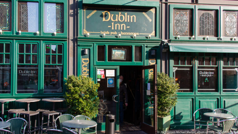 Green Dublin Inn pub façade with gold lettering, paneled windows, doorway, outdoor tables, and potted shrubs.