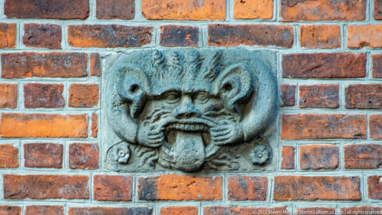 Weathered gray stone grotesque with a protruding tongue mounted in a red-brick wall.