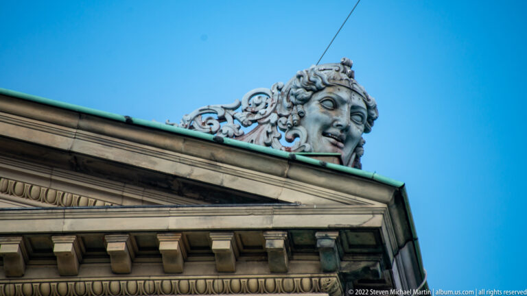Large sculpted female face with curling ornament rising above the opera house cornice against blue sky.