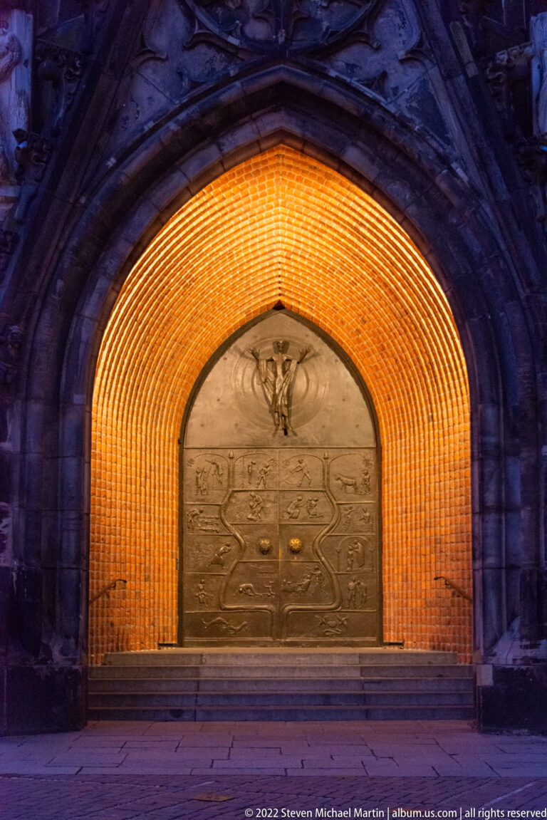 Large bronze relief doors inside a pointed brick arch glowing with warm amber lighting at night.