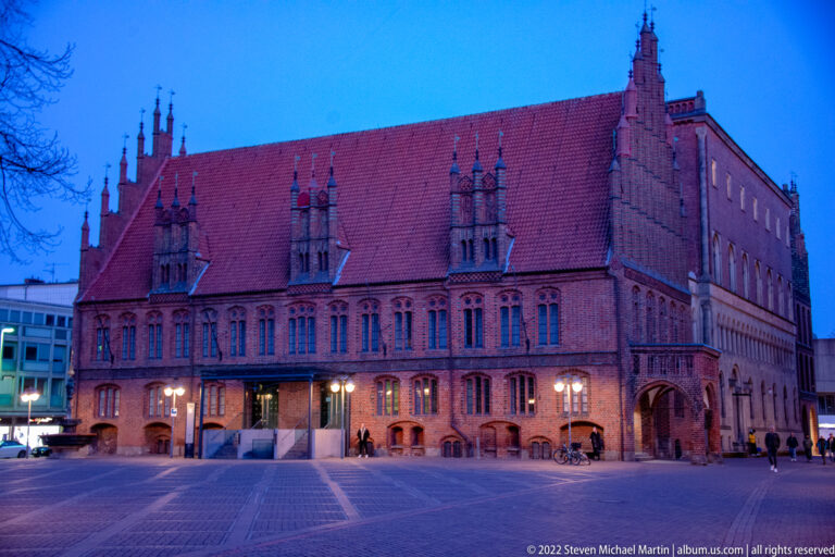 Long red-brick Gothic hall with steep roof and stepped gables illuminated by streetlamps under a blue evening sky.