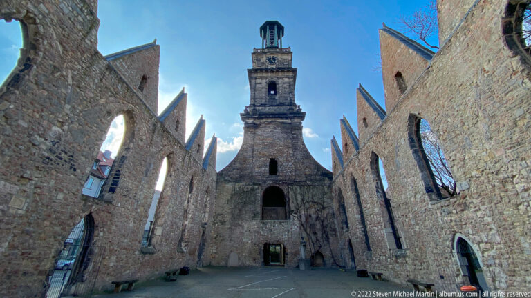 Roofless Aegidienkirche interior with tall ruined walls, open window arches, and the surviving tower under blue sky.
