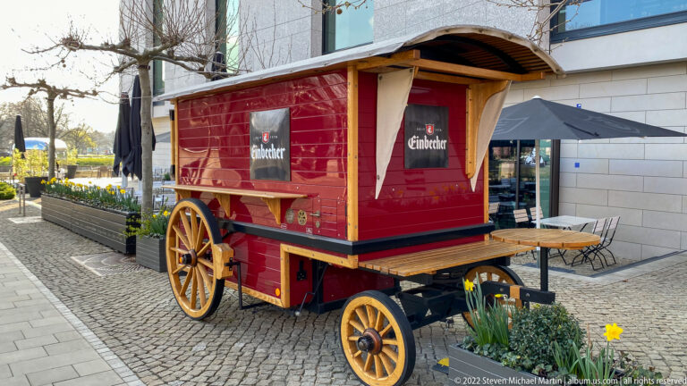 Red Einbecker beer wagon with wooden-spoked wheels, curved roof, and small serving shelves beside outdoor seating.