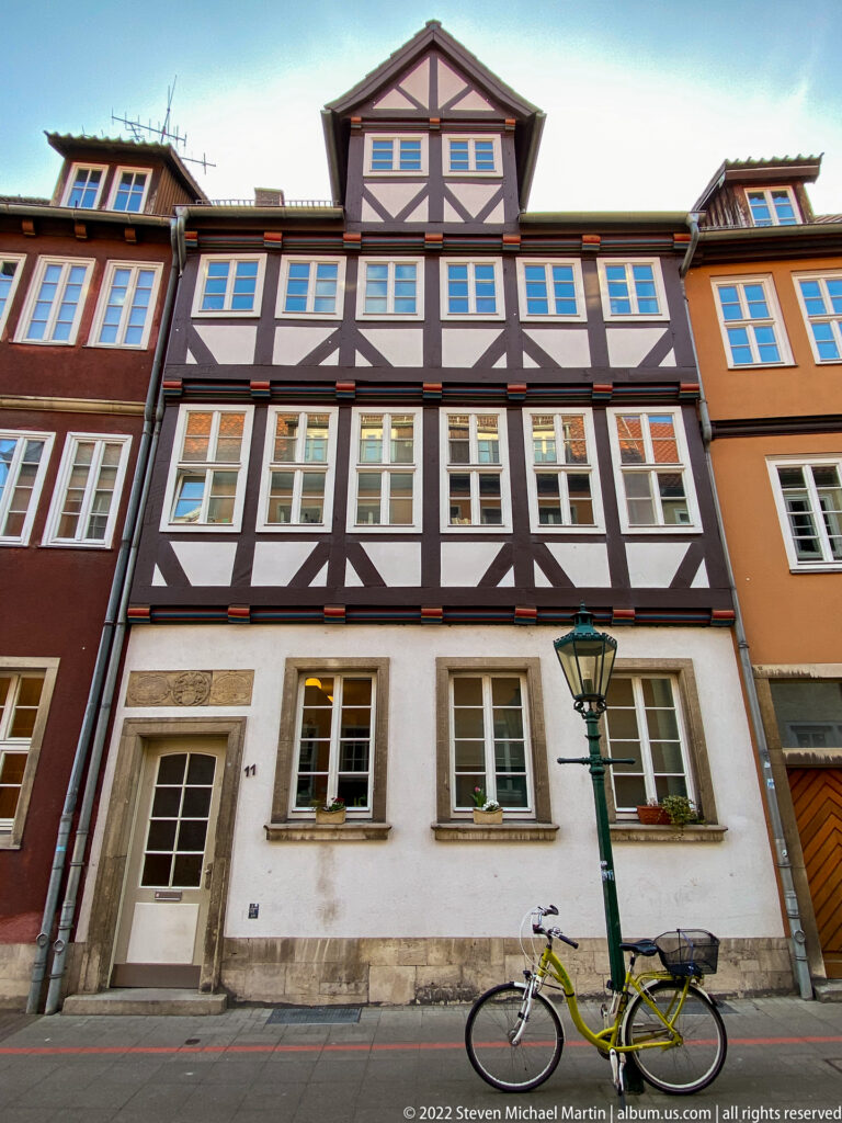Half-timbered townhouse with dark framing, white windows, pale ground floor, bicycle, and traditional streetlamp.