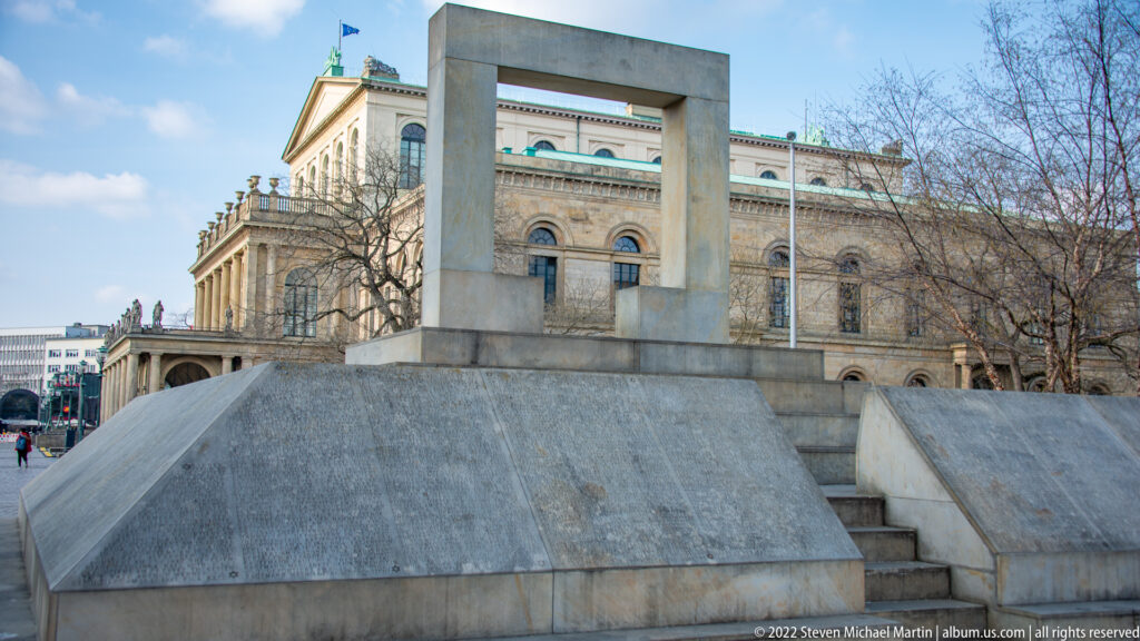 Modern pale stone memorial with rectangular opening and sloping base in front of the historic opera house.