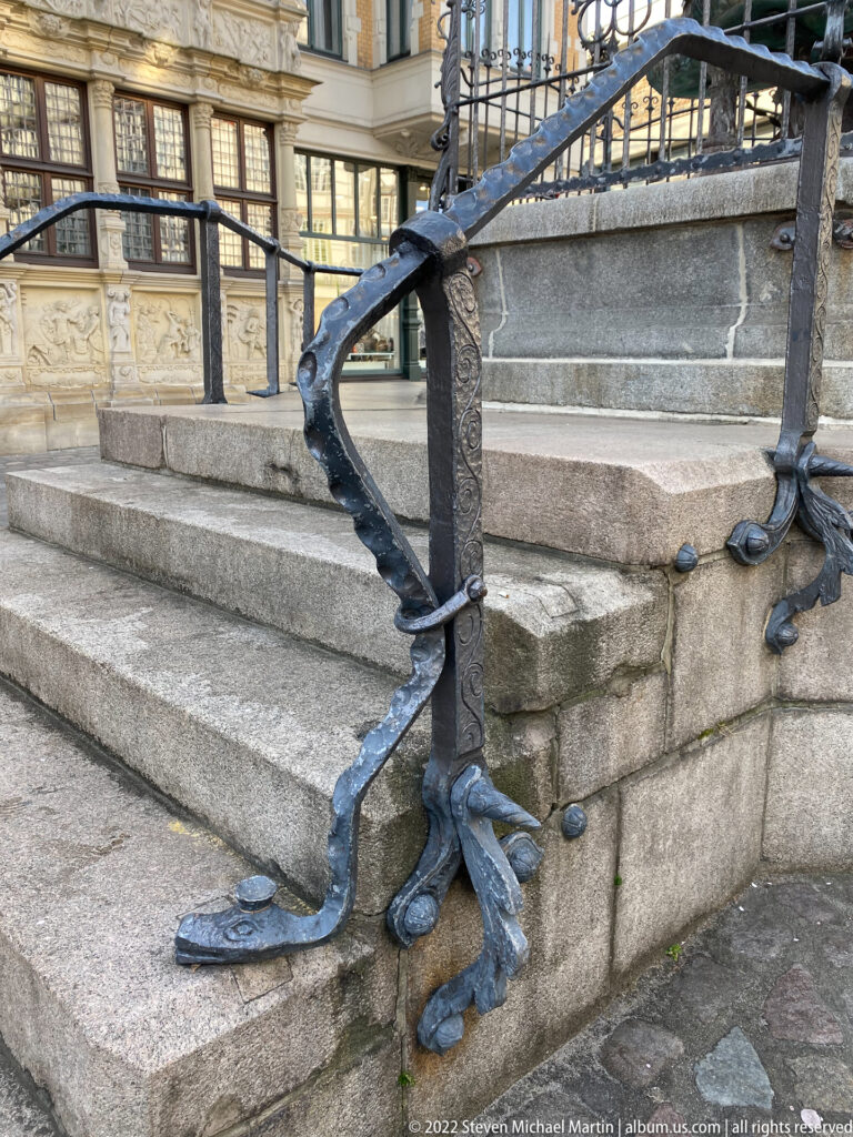 Forged metal stair railing with curling, root-like feet spreading across gray stone steps.