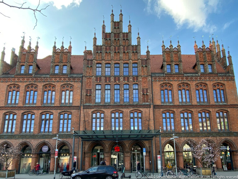 Ornate red-brick civic façade with stepped gables, pinnacles, red-tiled roof, arcaded ground floor, shops, bicycles, and cars.