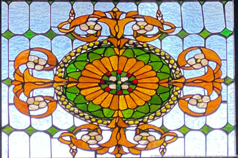 Stained-glass panel with orange-and-green central flower, curling stems, pale blossoms, green diamonds, and textured translucent background.