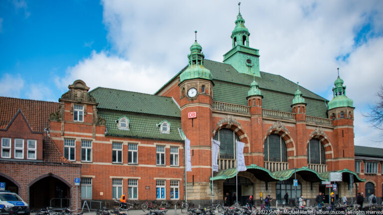 Red-brick Lübeck Hauptbahnhof with arched entrances, clock tower, green copper roofs, bicycles, and station canopy.