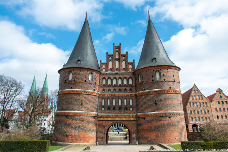 Frontal view of the Holstentor, showing twin round towers, dark conical roofs, brick bands, and central arched passage.