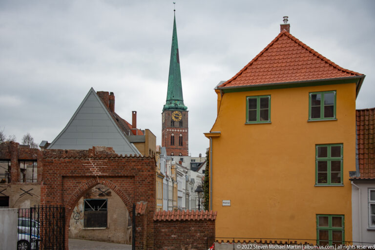 Green-spired brick church tower rising behind old brick walls, gabled roofs, and an ochre-colored house.