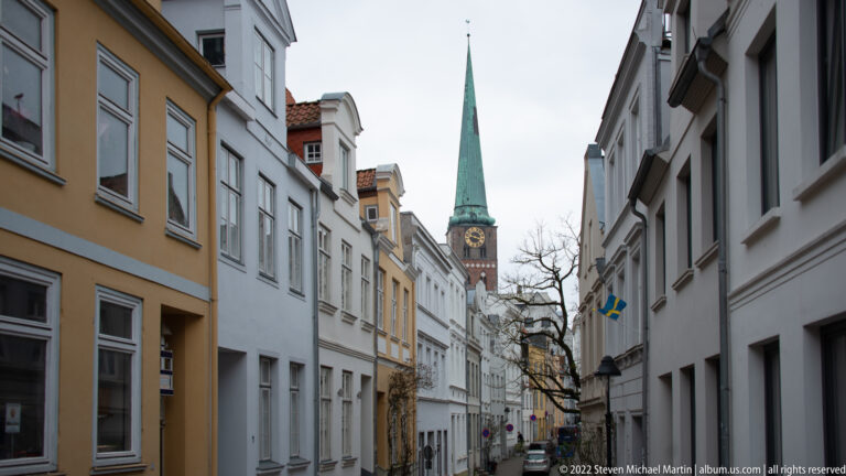 Narrow street lined with pale historic façades leading toward a green-spired brick church tower.