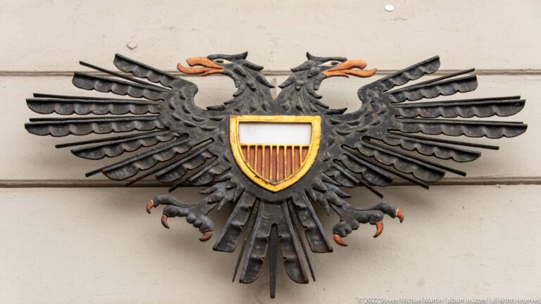 Black double-headed eagle with orange beaks and talons carrying a small red-and-white shield on a pale wall.