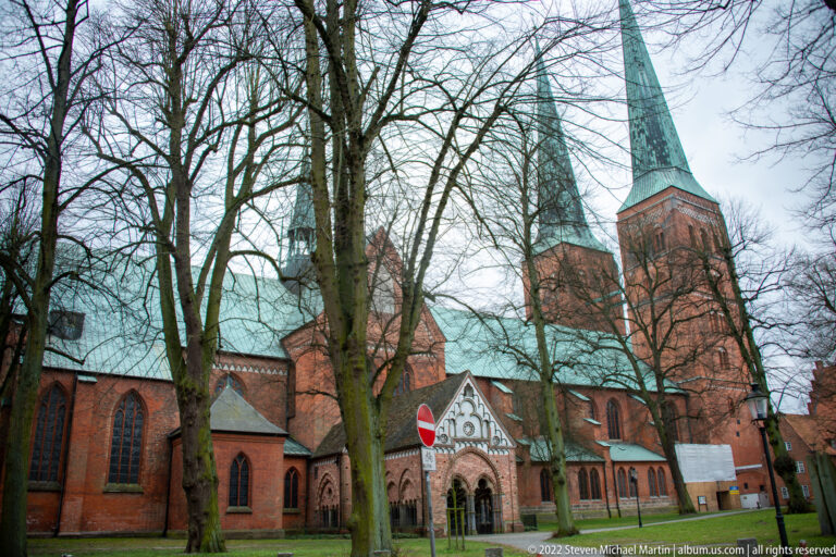 Long red-brick Gothic church with tall green spire and pointed windows, viewed through leafless trees across a lawn.