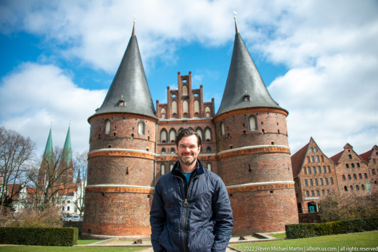Man in a dark jacket standing on grass before the twin brick towers and central gateway of the Holstentor.