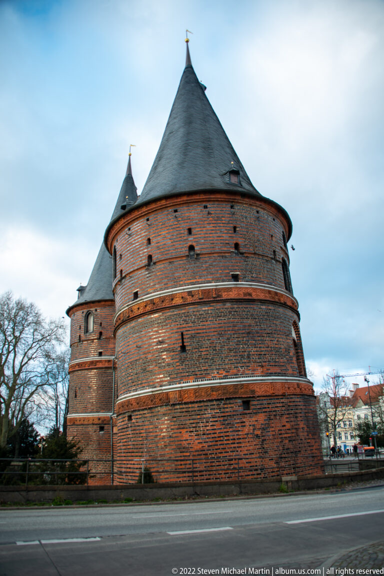 Large round brick defensive tower with very small openings and a broad dark conical roof beside a road.