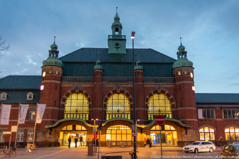 Lübeck Hauptbahnhof at dusk with three illuminated arched entrances, brick towers, cupolas, and central clock.