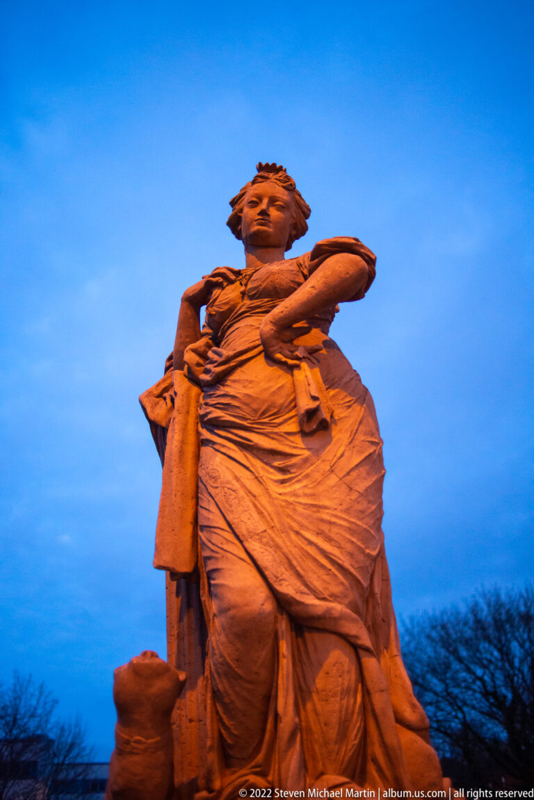Large illuminated female sculpture in layered garments viewed from below against a deep blue evening sky.
