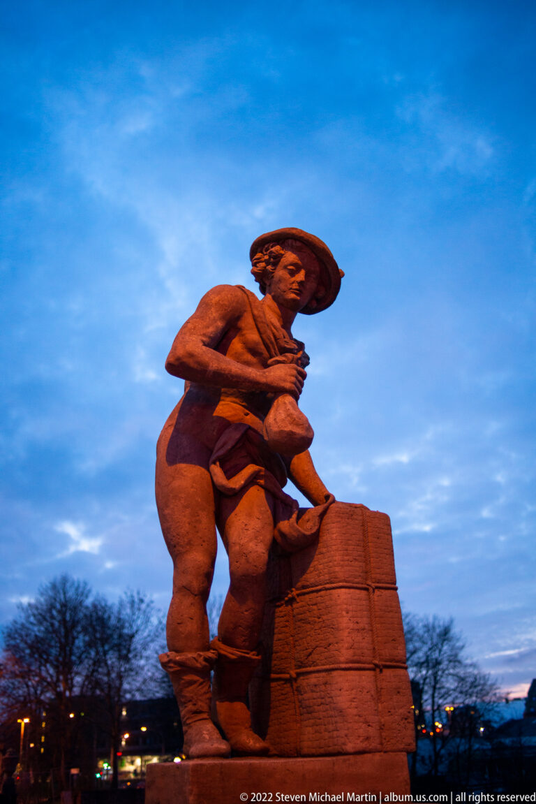 Large illuminated male sculpture with broad hat and heavy pack leaning over a block-like form at dusk.