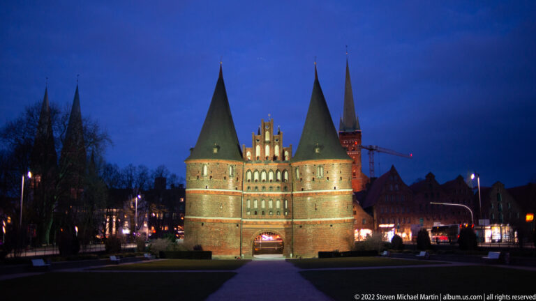 Illuminated Holstentor at blue hour with twin round towers, steep roofs, central gable, and arched passage.