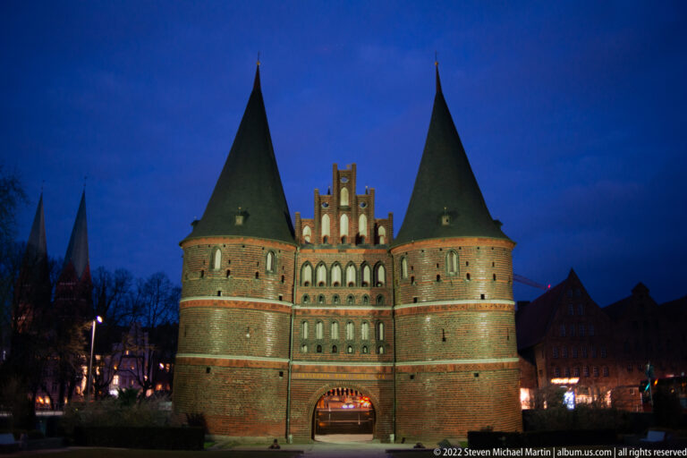 Closer night view of the Holstentor with floodlit brickwork, twin conical towers, stepped gable, and central arch.