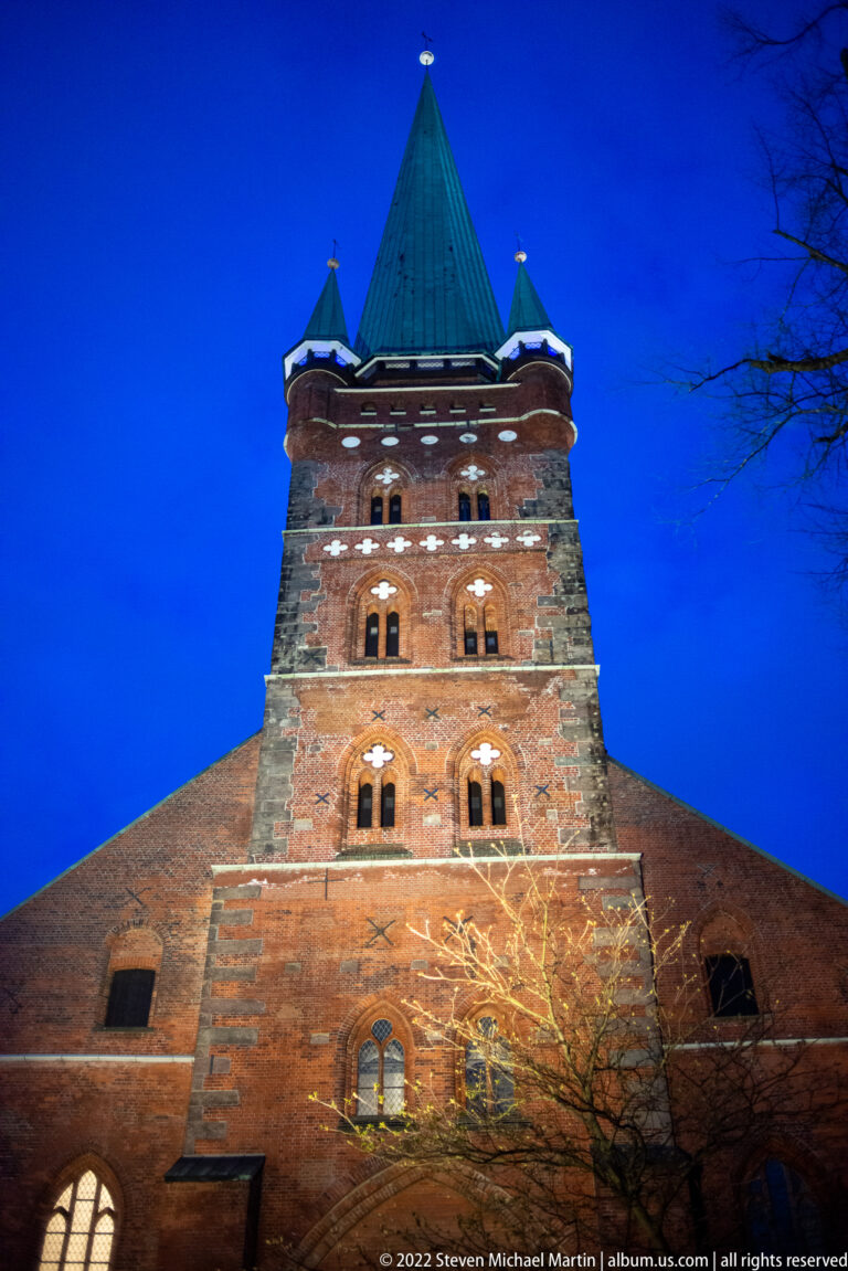 Illuminated west tower of St. Petri in Lübeck, with red-brick masonry, tall green copper spire, and four corner turrets at blue hour.