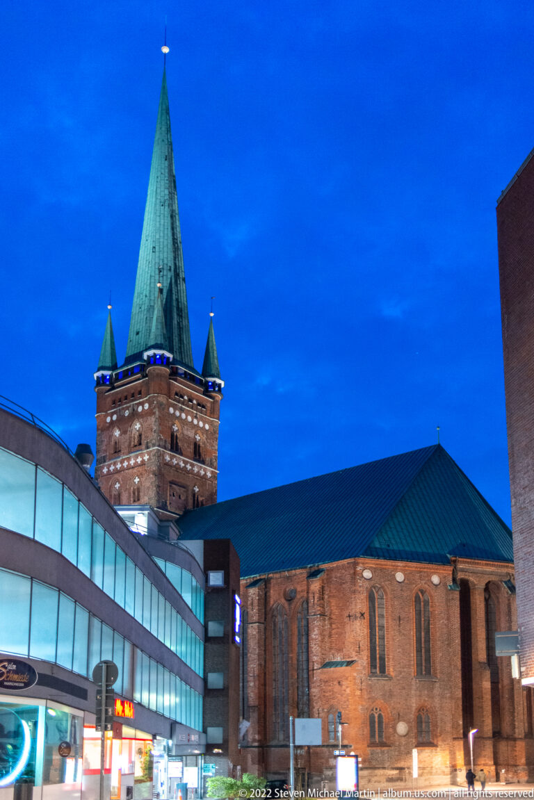 St. Petri Church in Lübeck at blue hour, showing its tall green copper spire, corner turrets, red-brick nave, and neighboring modern buildings.